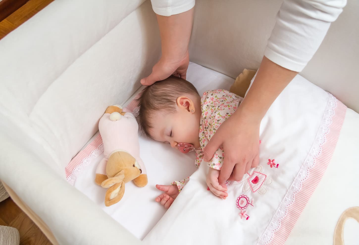 An image of a mother's hands caressing her cute baby girl sleeping in a cot with a pacifier and stuffed toy.
