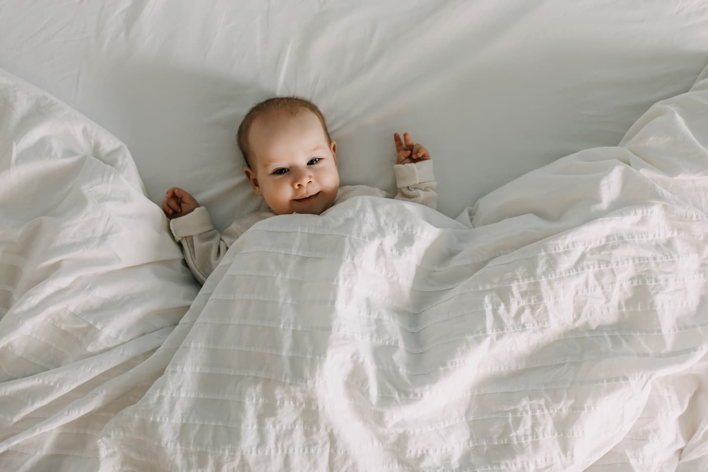 An image of a Happy baby lying in bed under a white soft blanket, smiling.