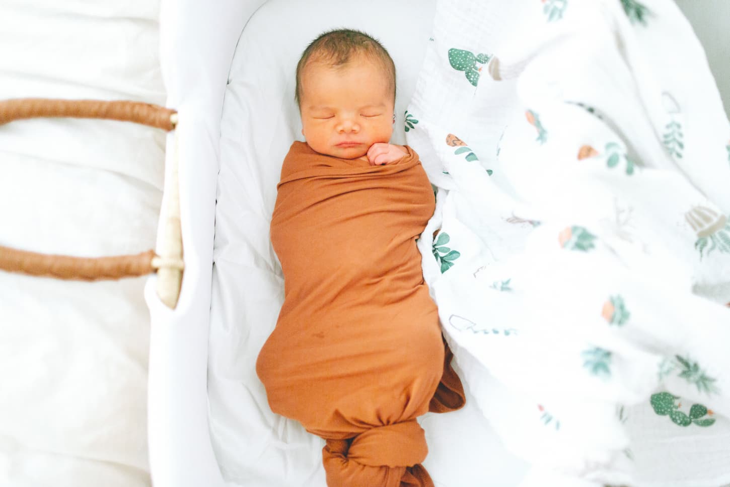 An image of a sleeping newborn baby swaddled in his bassinet in an earth-tone swaddling blanket.