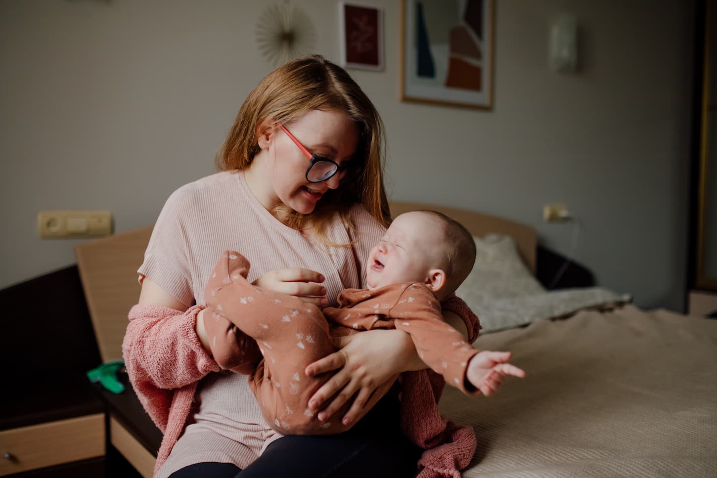An image of a sitting mother holding a 6-8 months old crying baby in his onesie in the bedroom.