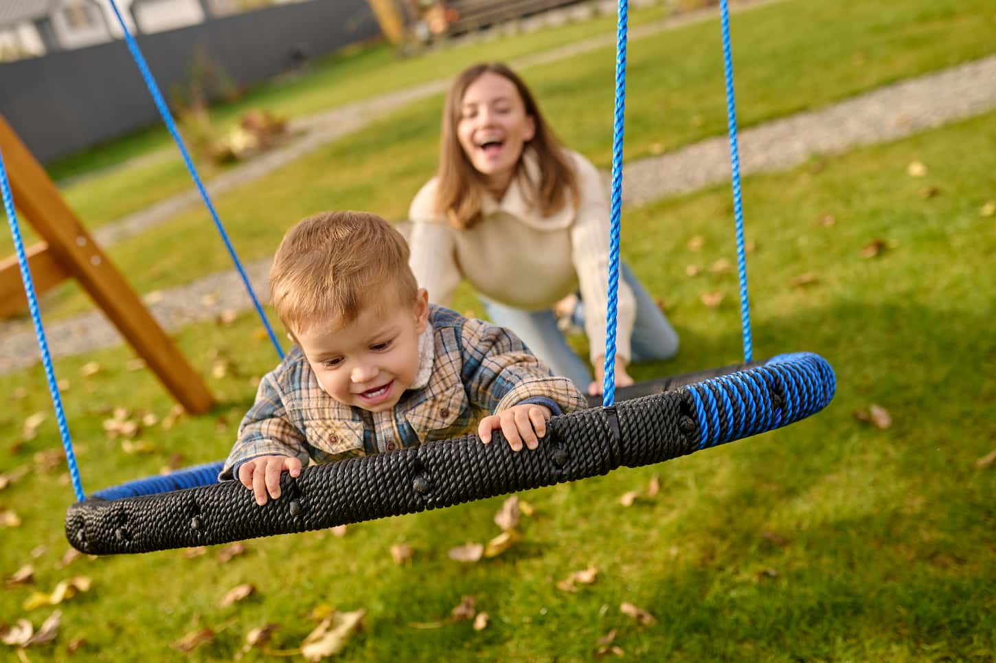An image of a Young enthusiastic blonde woman in comfortable casual clothes kneeling pushing a swing with a joyful child on a green lawn on a sunny autumn day.