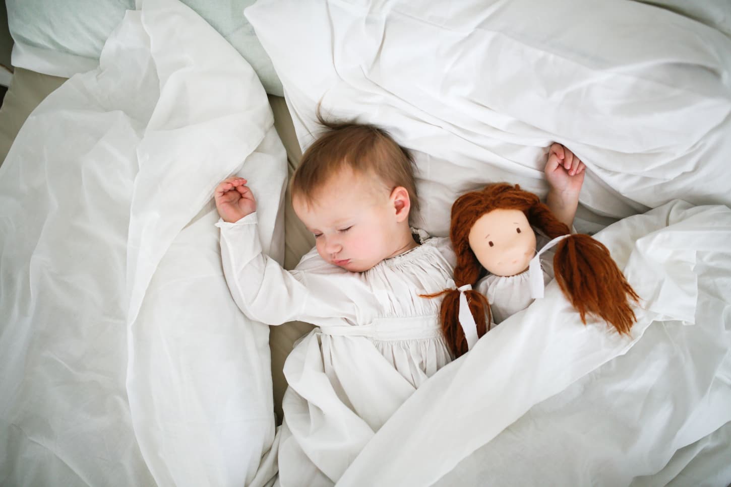 An image of a sick baby lying in bed with a doll beside her while having bed rest during illness.