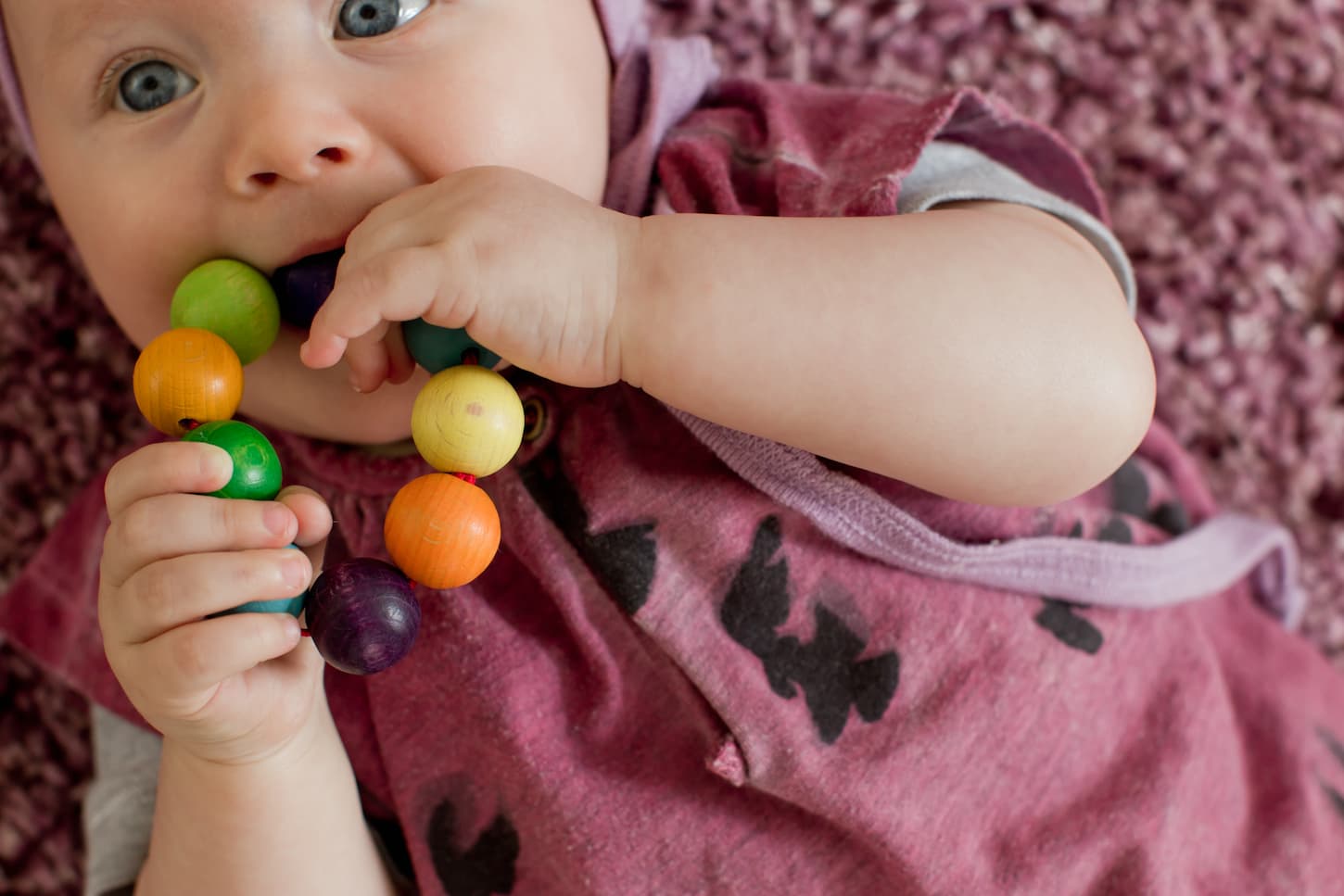 An image of a Baby girl wearing a pink bodysuit with a teething toy in her mouth.