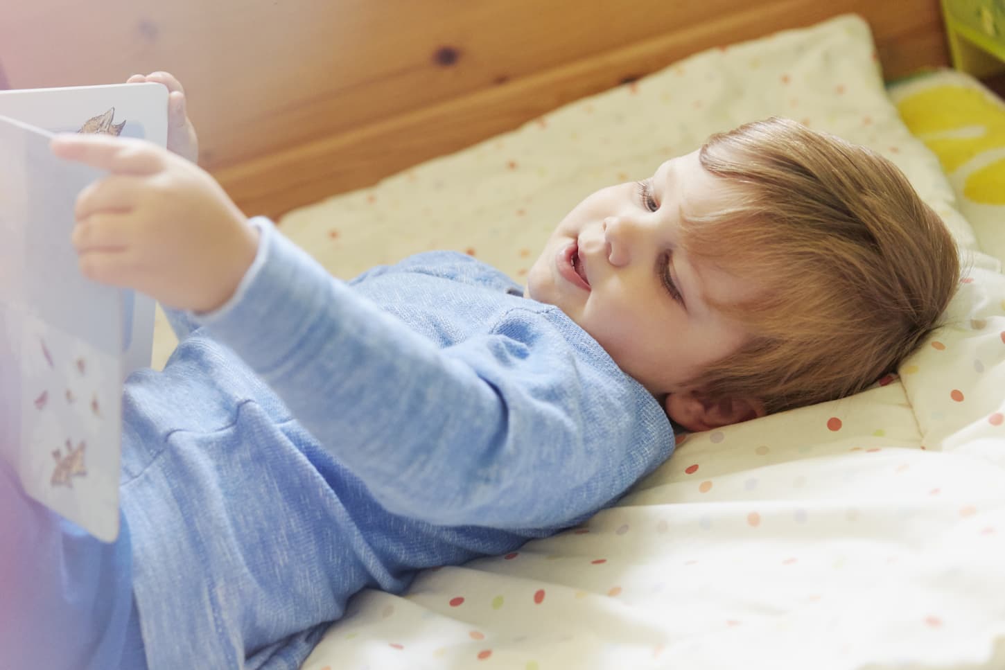 An image of a Boy lying on his back in bed looking at a storybook.
