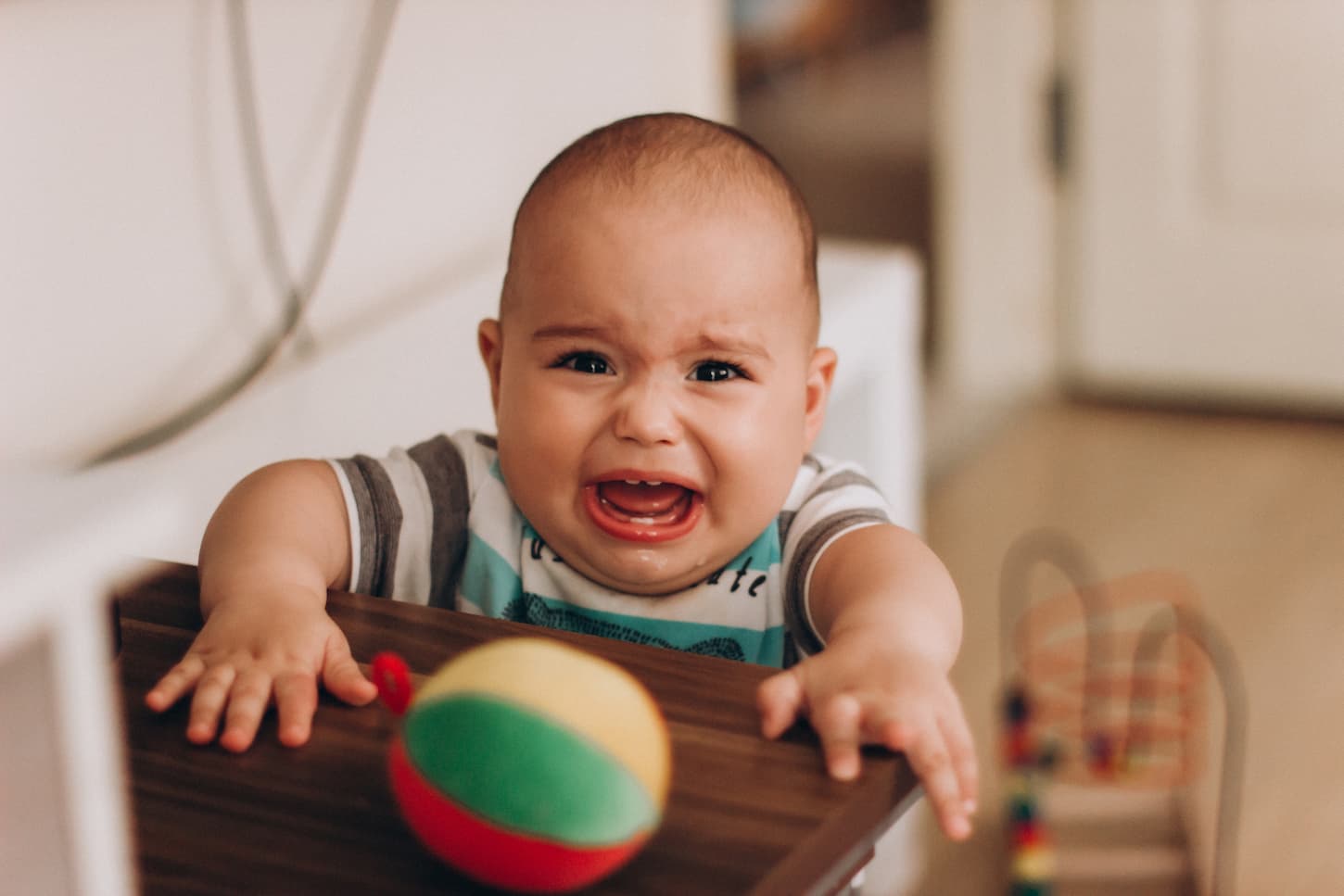 An image of a Crying little baby boy standing near a table reaching for his toy.