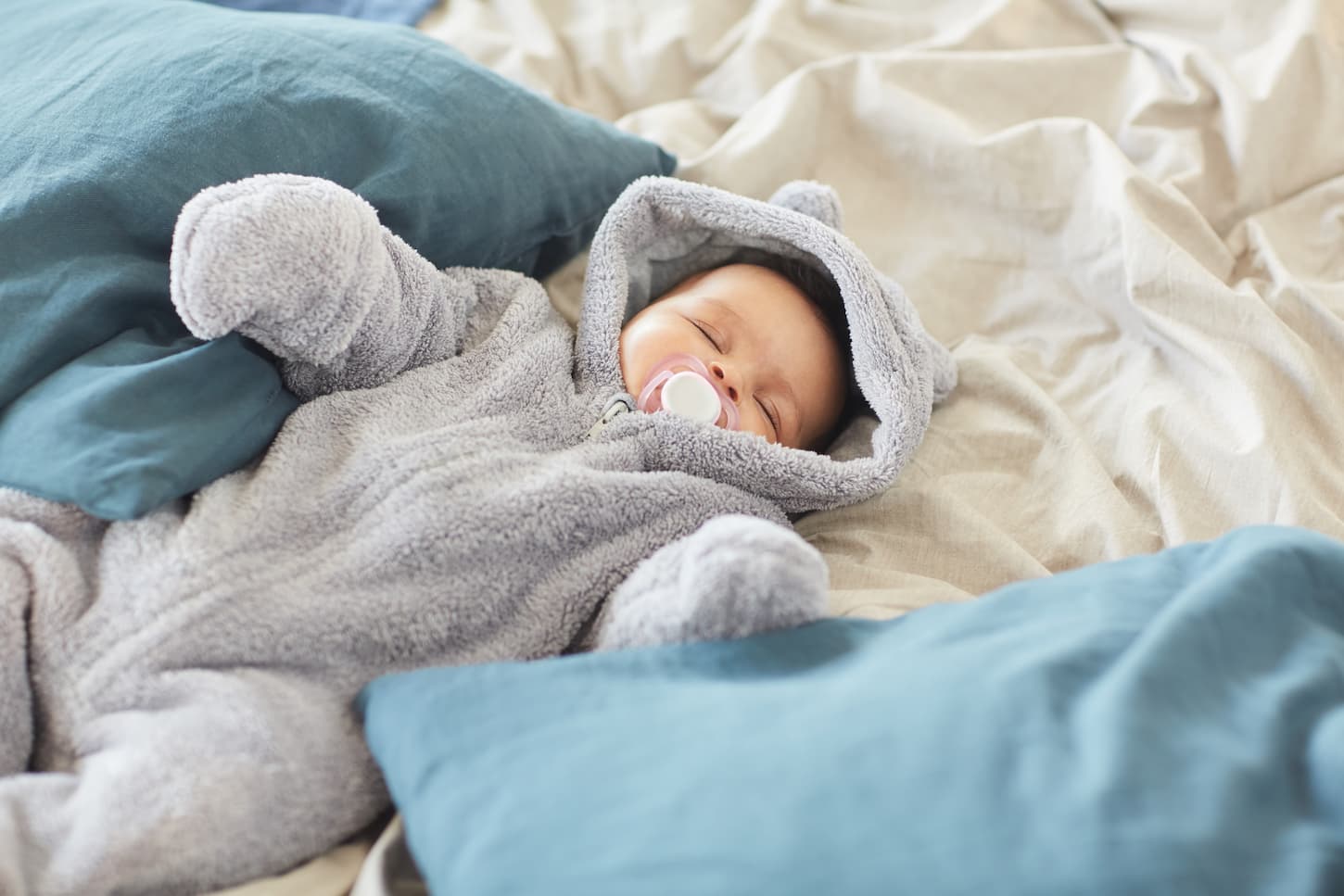 An image of a cute baby wearing a fluffy fleece onesie sucking on a pacifier while sleeping lying on bed blankets.