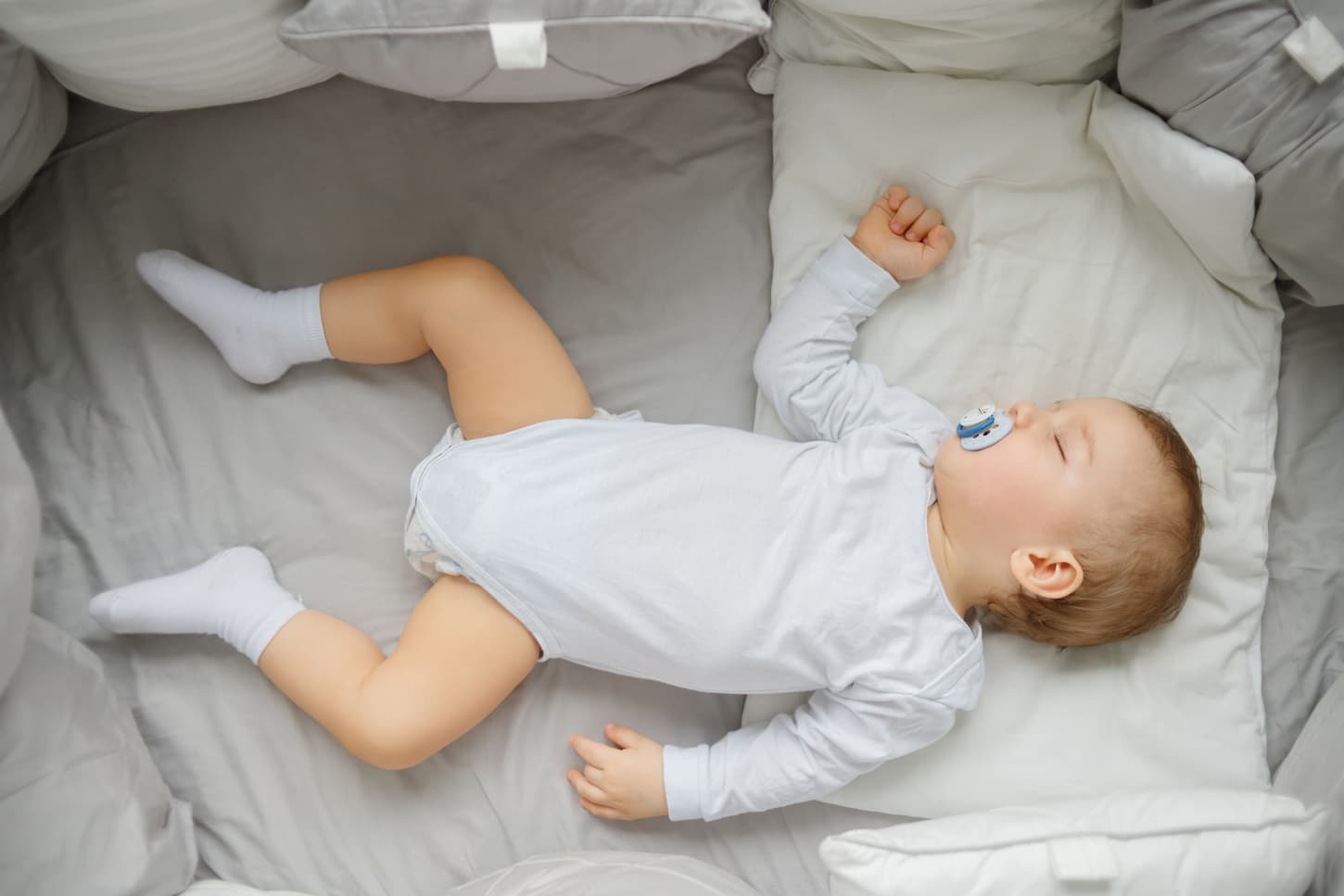 An image of a Little baby sleeping in a crib with a pacifier on and wearing his bodysuit.