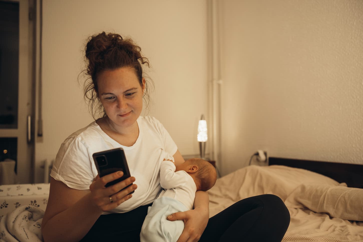 An image of a Young busy mother using a smartphone and breastfeeding her baby boy while sitting on a bed at home.