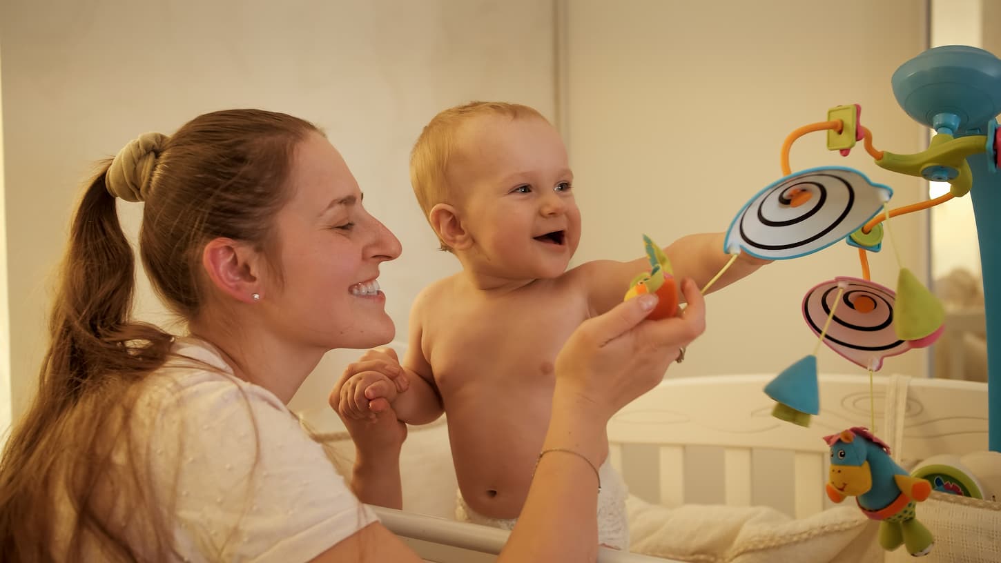 An image of a smiling baby with his mother playing with spinning toys hanging on mobile in bed at night.